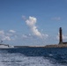 A white naval or coast guard patrol vessel is navigating past a harbor entrance marked by a red brick lighthouse mounted on a stone breakwater. The scene appears to be a port entrance with calm blue waters and a partly cloudy sky. The vessel appears to be either departing or passing the harbor mouth.