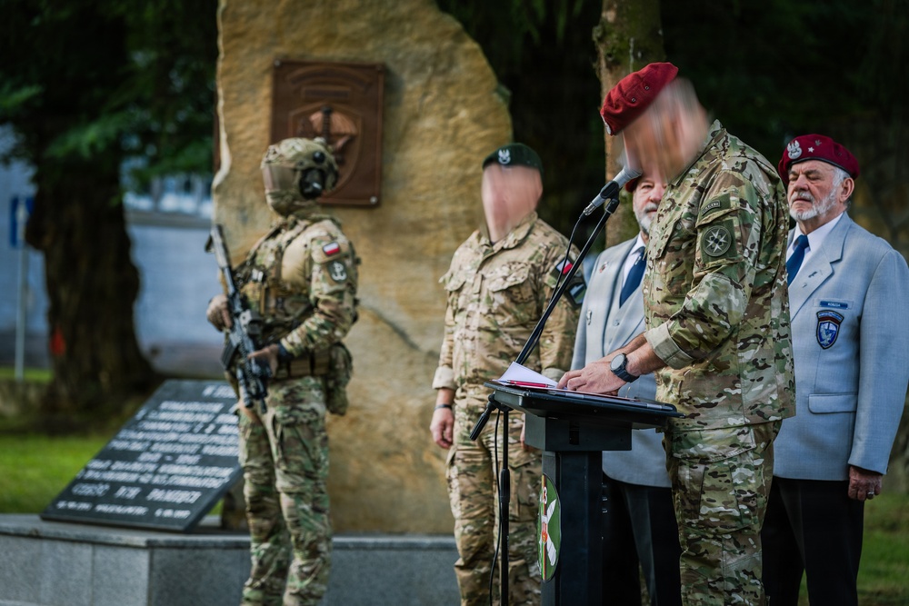 A military ceremony is taking place in front of a stone memorial monument with a bronze plaque. Several personnel in camouflage uniforms are present, including one speaking at a podium, while an armed soldier in full tactical gear stands guard. A civilian elder in a light gray jacket with a unit badge is also present, suggesting a commemorative or memorial event.