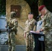 A military ceremony is taking place in front of a stone memorial monument with a bronze plaque. Several personnel in camouflage uniforms are present, including one speaking at a podium, while an armed soldier in full tactical gear stands guard. A civilian elder in a light gray jacket with a unit badge is also present, suggesting a commemorative or memorial event.