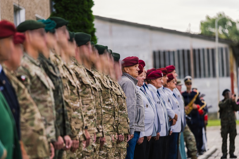 A military parade or ceremonial formation featuring active-duty soldiers in camouflage uniforms alongside elderly veterans wearing red/maroon berets and civilian dress with medals. The formation is standing at attention outdoors near a building, suggesting a commemorative or official military ceremony. A military band with brass instruments is visible in the background.