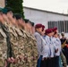 A military parade or ceremonial formation featuring active-duty soldiers in camouflage uniforms alongside elderly veterans wearing red/maroon berets and civilian dress with medals. The formation is standing at attention outdoors near a building, suggesting a commemorative or official military ceremony. A military band with brass instruments is visible in the background.