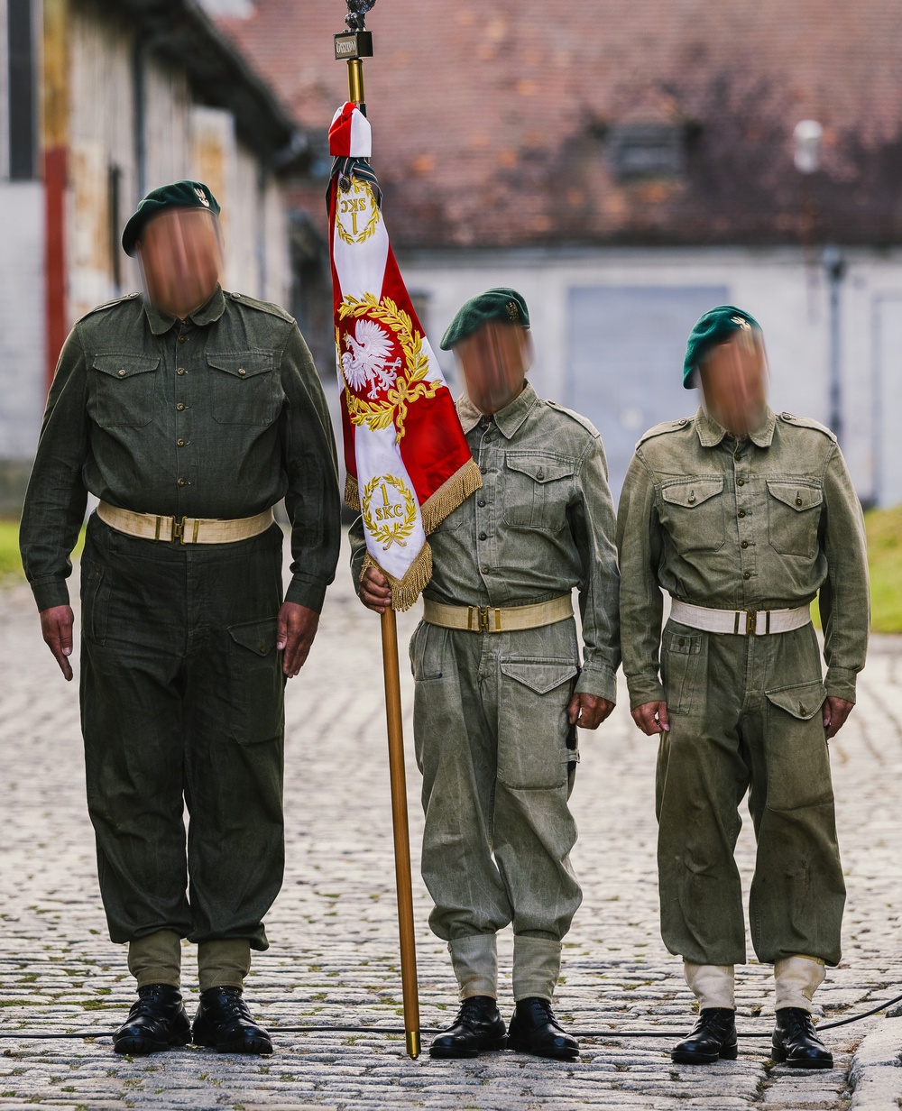Three individuals in military-style uniforms stand on a cobblestone surface, with the central figure holding a ceremonial flag/standard. The flag displays red and white colors with a Polish eagle emblem and gold embroidery, along with the inscription '1 SKC'. The individuals wear green berets and olive-drab field uniforms with white belts, suggesting a formal military or reenactment ceremony.