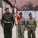 Three individuals in military-style uniforms stand on a cobblestone surface, with the central figure holding a ceremonial flag/standard. The flag displays red and white colors with a Polish eagle emblem and gold embroidery, along with the inscription '1 SKC'. The individuals wear green berets and olive-drab field uniforms with white belts, suggesting a formal military or reenactment ceremony.