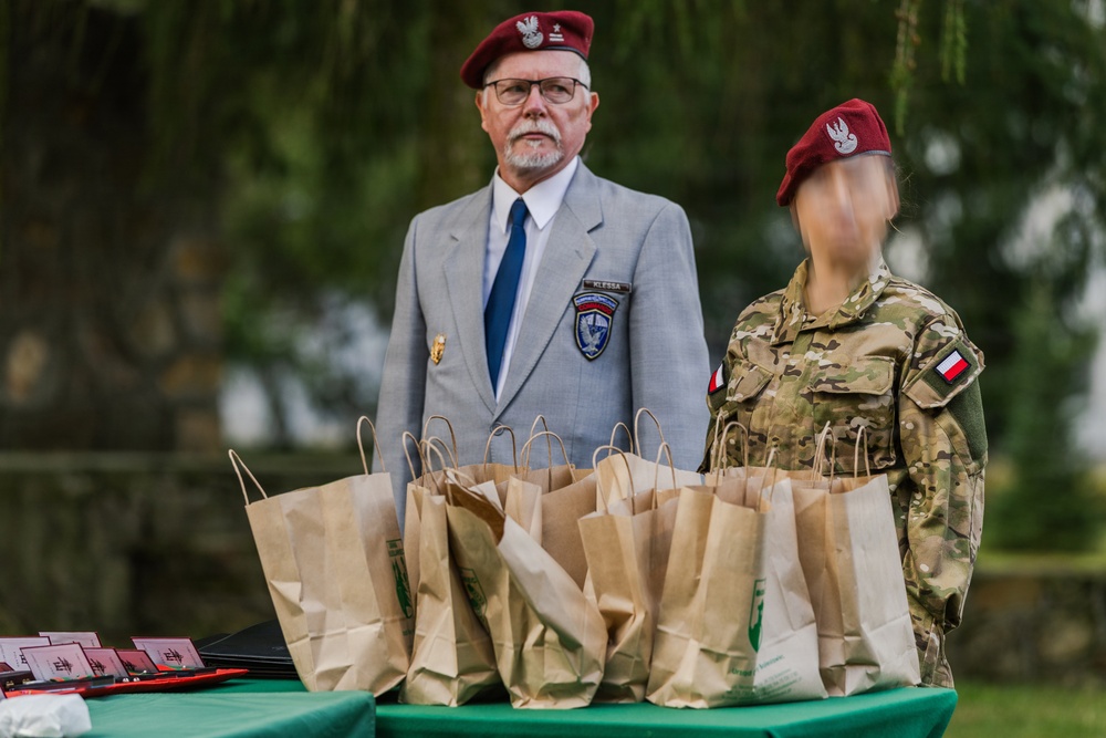 Two individuals wearing matching dark red/maroon berets stand behind a table covered with numerous brown paper bags at what appears to be an outdoor military or veterans' ceremony. The older man on the left wears a gray blazer with a unit patch and name tag, while the person on the right wears a Polish military multicam uniform with a Polish flag patch. The table appears to be set up for a distribution or award event.