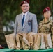Two individuals wearing matching dark red/maroon berets stand behind a table covered with numerous brown paper bags at what appears to be an outdoor military or veterans' ceremony. The older man on the left wears a gray blazer with a unit patch and name tag, while the person on the right wears a Polish military multicam uniform with a Polish flag patch. The table appears to be set up for a distribution or award event.