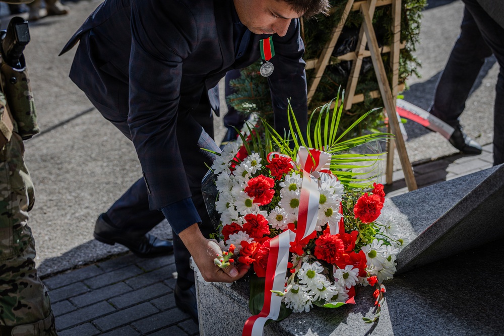 A man wearing a dark suit and a medal is laying a floral wreath on a stone monument or memorial. The wreath consists of red carnations and white chrysanthemums adorned with a red-and-white ribbon, suggesting a formal commemorative ceremony. Military personnel in camouflage uniforms are visible in the background, indicating an official state or military memorial event.