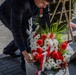 A man wearing a dark suit and a medal is laying a floral wreath on a stone monument or memorial. The wreath consists of red carnations and white chrysanthemums adorned with a red-and-white ribbon, suggesting a formal commemorative ceremony. Military personnel in camouflage uniforms are visible in the background, indicating an official state or military memorial event.