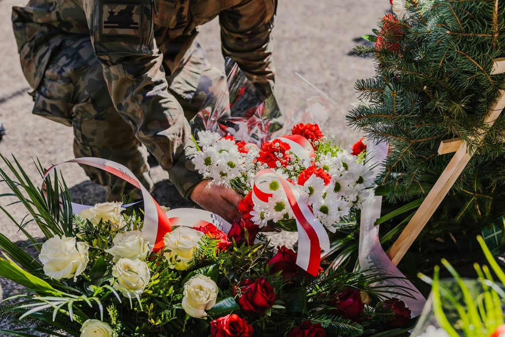 A person in military camouflage uniform is placing or arranging floral wreaths, likely at a memorial or ceremonial site. The wreaths are decorated with red and white ribbons, consistent with Polish national colors. The scene appears to be part of a formal commemorative ceremony.