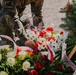 A person in military camouflage uniform is placing or arranging floral wreaths, likely at a memorial or ceremonial site. The wreaths are decorated with red and white ribbons, consistent with Polish national colors. The scene appears to be part of a formal commemorative ceremony.
