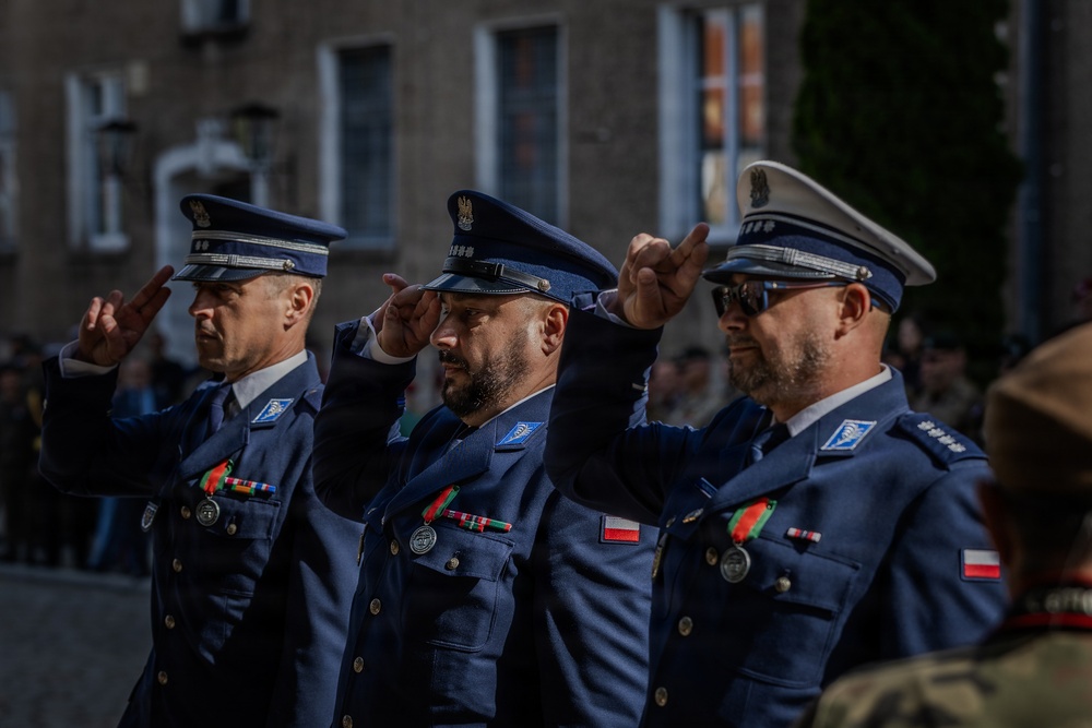 Three uniformed officers in blue dress uniforms are performing a formal salute during what appears to be an official ceremony. They wear peaked caps with eagle insignia and display medal ribbons and decorations on their chests, consistent with Polish military or law enforcement dress uniforms. A blurred crowd and historic building are visible in the background.