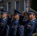 Three uniformed officers in blue dress uniforms are performing a formal salute during what appears to be an official ceremony. They wear peaked caps with eagle insignia and display medal ribbons and decorations on their chests, consistent with Polish military or law enforcement dress uniforms. A blurred crowd and historic building are visible in the background.
