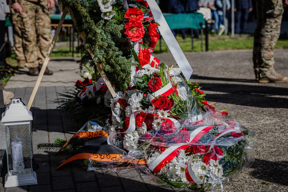 A memorial ceremony is taking place outdoors, featuring multiple floral wreaths laid on a paved surface. The wreaths are decorated with red and white ribbons consistent with Polish national colors, along with an orange and black ribbon visible on one arrangement. Military personnel in camouflage uniforms are visible in the background, suggesting an official commemorative event.