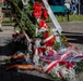 A memorial ceremony is taking place outdoors, featuring multiple floral wreaths laid on a paved surface. The wreaths are decorated with red and white ribbons consistent with Polish national colors, along with an orange and black ribbon visible on one arrangement. Military personnel in camouflage uniforms are visible in the background, suggesting an official commemorative event.