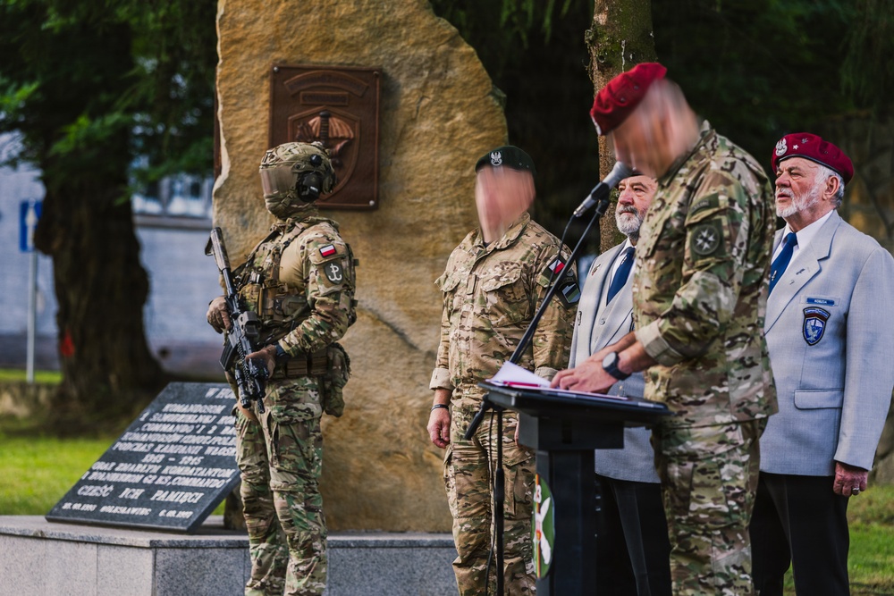 A military ceremony is taking place in front of a stone memorial monument with a bronze plaque. Several individuals in military uniforms and civilian attire are gathered, with one person speaking at a podium/lectern. The setting appears to be an outdoor commemorative event, likely in Poland based on visible Polish flag patches and text on the monument.