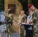 A military ceremony is taking place in front of a stone memorial monument with a bronze plaque. Several individuals in military uniforms and civilian attire are gathered, with one person speaking at a podium/lectern. The setting appears to be an outdoor commemorative event, likely in Poland based on visible Polish flag patches and text on the monument.
