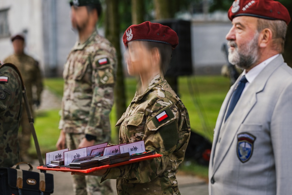 A military ceremony appears to be taking place, likely in Poland based on the visible Polish flag patches on uniforms. A soldier in camouflage uniform and maroon beret is carrying a red tray with what appear to be medals or awards in presentation boxes. An older civilian man in a gray suit wearing a maroon beret and organizational badge stands nearby.