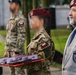 A military ceremony appears to be taking place, likely in Poland based on the visible Polish flag patches on uniforms. A soldier in camouflage uniform and maroon beret is carrying a red tray with what appear to be medals or awards in presentation boxes. An older civilian man in a gray suit wearing a maroon beret and organizational badge stands nearby.
