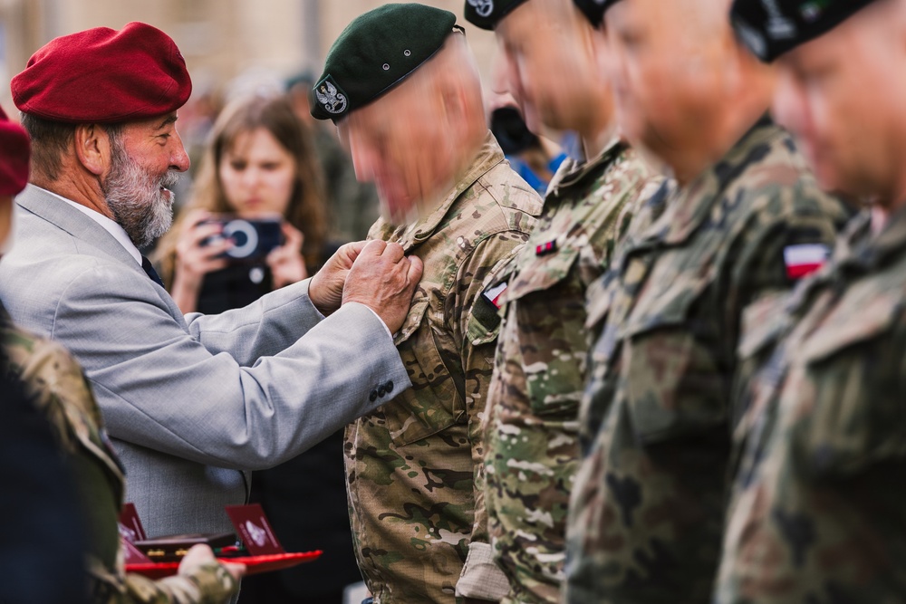 A military decoration ceremony is taking place outdoors, where an older civilian man wearing a red beret and gray suit is pinning a medal or badge onto a soldier in camouflage uniform. A line of soldiers wearing green and dark berets with Polish flag patches on their sleeves stand at attention. A civilian woman is visible in the background photographing the event.