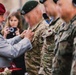 A military decoration ceremony is taking place outdoors, where an older civilian man wearing a red beret and gray suit is pinning a medal or badge onto a soldier in camouflage uniform. A line of soldiers wearing green and dark berets with Polish flag patches on their sleeves stand at attention. A civilian woman is visible in the background photographing the event.