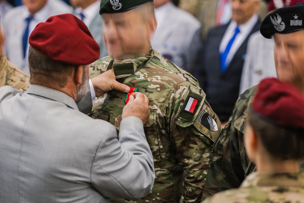 A military decoration ceremony is taking place where a civilian or senior official in a gray suit and red beret is pinning a medal or ribbon onto a soldier in multicam camouflage uniform. The soldier wears a green beret and displays a French flag patch along with a 'Commando' unit patch on the left sleeve. Several other military personnel wearing red and black berets are visible in the background.