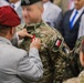 A military decoration ceremony is taking place where a civilian or senior official in a gray suit and red beret is pinning a medal or ribbon onto a soldier in multicam camouflage uniform. The soldier wears a green beret and displays a French flag patch along with a 'Commando' unit patch on the left sleeve. Several other military personnel wearing red and black berets are visible in the background.