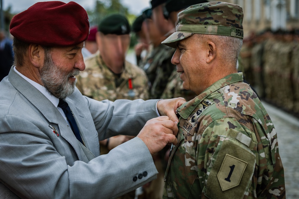 A civilian man wearing a red beret and gray suit is pinning a medal or decoration onto a U.S. Army soldier in OCP (Operational Camouflage Pattern) uniform during what appears to be a formal military ceremony. The soldier's uniform displays a '1' patch (1st Infantry Division) and a name tape reading 'DEMO' or similar. Multiple military personnel in various uniforms are visible in the background.