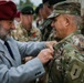 A civilian man wearing a red beret and gray suit is pinning a medal or decoration onto a U.S. Army soldier in OCP (Operational Camouflage Pattern) uniform during what appears to be a formal military ceremony. The soldier's uniform displays a '1' patch (1st Infantry Division) and a name tape reading 'DEMO' or similar. Multiple military personnel in various uniforms are visible in the background.