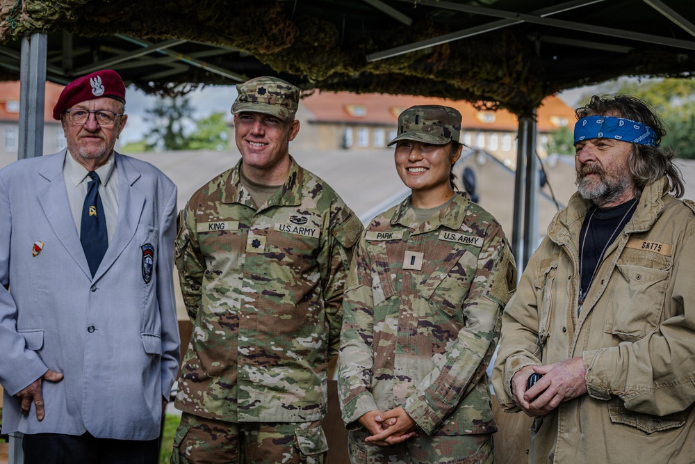 Four individuals pose together under a camouflage netting canopy, likely at a military event or ceremony. Two are active U.S. Army personnel in OCP (Operational Camouflage Pattern) uniforms identified by name tapes as 'KING' and 'PARK', while a third wears a red beret and civilian attire suggesting veteran status, and a fourth is a civilian in casual clothing. The setting appears to be an outdoor military installation with brick buildings visible in the background.