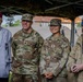 Four individuals pose together under a camouflage netting canopy, likely at a military event or ceremony. Two are active U.S. Army personnel in OCP (Operational Camouflage Pattern) uniforms identified by name tapes as 'KING' and 'PARK', while a third wears a red beret and civilian attire suggesting veteran status, and a fourth is a civilian in casual clothing. The setting appears to be an outdoor military installation with brick buildings visible in the background.