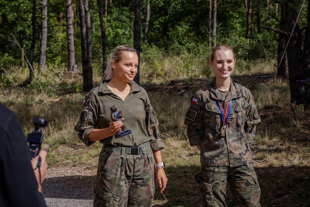Two female soldiers in camouflage uniforms stand outdoors in a forested area, appearing to be at an award or recognition ceremony. The soldier on the left holds a small trophy or figurine, while the soldier on the right wears a medal on a ribbon around her neck. A photographer or videographer is partially visible in the foreground left.