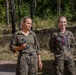 Two female soldiers in camouflage uniforms stand outdoors in a forested area, appearing to be at an award or recognition ceremony. The soldier on the left holds a small trophy or figurine, while the soldier on the right wears a medal on a ribbon around her neck. A photographer or videographer is partially visible in the foreground left.