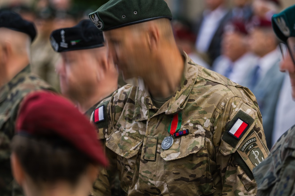 A military ceremony featuring soldiers in Polish Armed Forces uniforms, with the central figure wearing multicam camouflage and displaying a medal with a red and green ribbon. The soldier's face is motion-blurred, likely due to movement during the shot. Multiple military personnel with various berets and insignia are visible in the background.