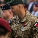 A military ceremony featuring soldiers in Polish Armed Forces uniforms, with the central figure wearing multicam camouflage and displaying a medal with a red and green ribbon. The soldier's face is motion-blurred, likely due to movement during the shot. Multiple military personnel with various berets and insignia are visible in the background.
