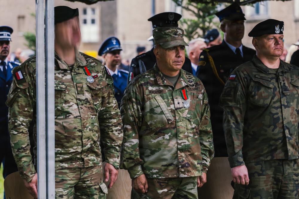 A military ceremony featuring personnel from multiple nations, including a clearly identified U.S. Army officer with the name tape 'DEMONTIONY' and two-star general rank insignia. Soldiers wearing camouflage uniforms with Polish and U.S. flags on their sleeves stand at attention alongside personnel in blue dress uniforms visible in the background. The setting appears to be an outdoor formal military event.