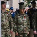 A military ceremony featuring personnel from multiple nations, including a clearly identified U.S. Army officer with the name tape 'DEMONTIONY' and two-star general rank insignia. Soldiers wearing camouflage uniforms with Polish and U.S. flags on their sleeves stand at attention alongside personnel in blue dress uniforms visible in the background. The setting appears to be an outdoor formal military event.