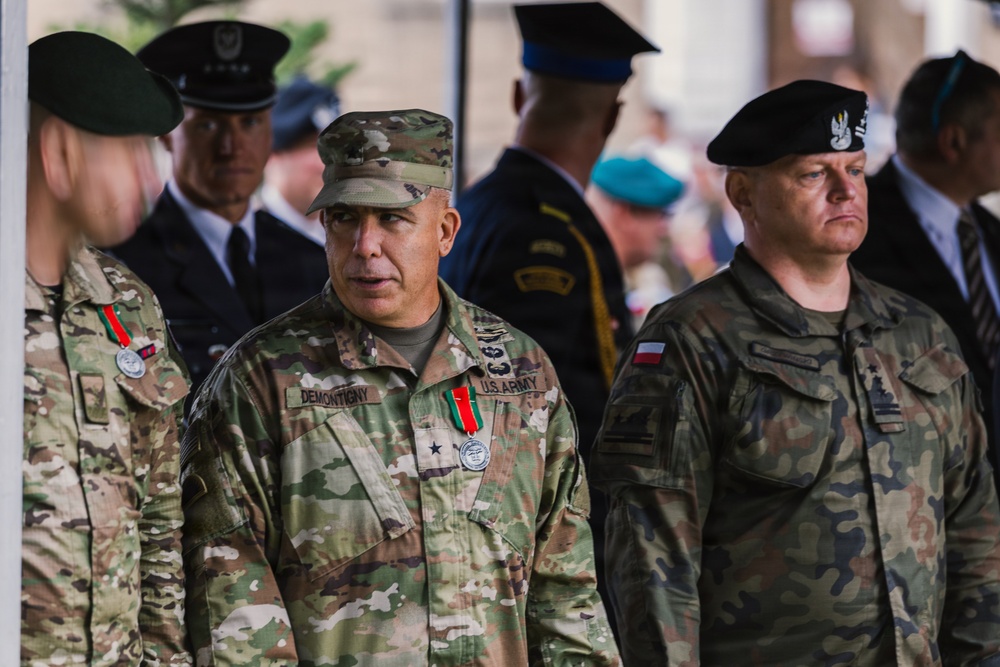 The image shows a multinational military gathering featuring U.S. Army and Polish Army personnel in uniform. A central U.S. Army officer with the name tape 'DEMONTIGNY' and a one-star general insignia is prominently visible. The setting appears to be a formal military ceremony or official event.