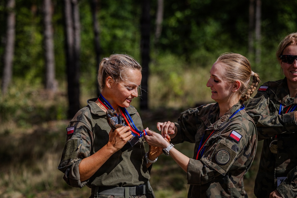 Two female military personnel in Polish Army camouflage uniforms are engaged in a medal presentation ceremony outdoors. One soldier is receiving or adjusting a medal on a red, white, and blue lanyard, while the other assists with placing it. A third uniformed individual is partially visible in the background.