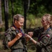 Two female military personnel in Polish Army camouflage uniforms are engaged in a medal presentation ceremony outdoors. One soldier is receiving or adjusting a medal on a red, white, and blue lanyard, while the other assists with placing it. A third uniformed individual is partially visible in the background.