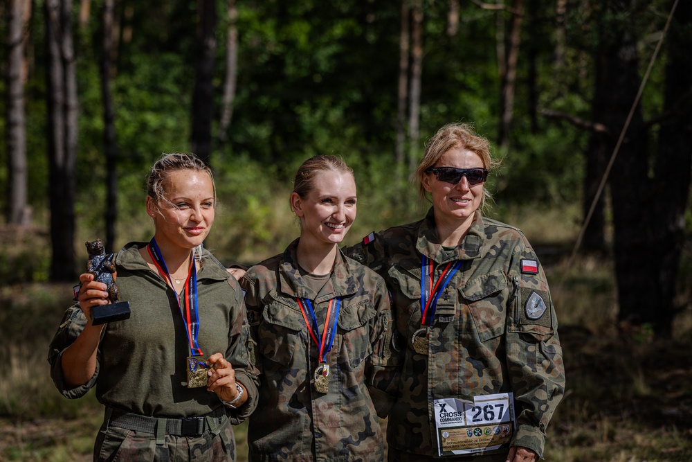 Three women in military camouflage uniforms pose together outdoors in a forested setting, displaying medals around their necks. One woman holds a trophy figurine while another wears a race bib numbered 267 from an event called 'Cross Commando.' They appear to be celebrating after completing a military competition or endurance event.