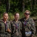 Three women in military camouflage uniforms pose together outdoors in a forested setting, displaying medals around their necks. One woman holds a trophy figurine while another wears a race bib numbered 267 from an event called 'Cross Commando.' They appear to be celebrating after completing a military competition or endurance event.