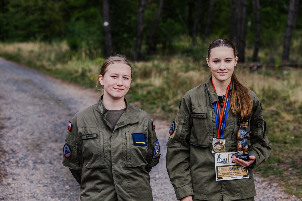 Two young women in matching olive/khaki military-style uniforms pose together on a gravel path surrounded by forest. The person on the right is wearing medals on lanyards and holding a trophy figurine, suggesting a recent competition award. Both uniforms feature Polish flag patches and unit insignia on the sleeves.
