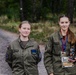 Two young women in matching olive/khaki military-style uniforms pose together on a gravel path surrounded by forest. The person on the right is wearing medals on lanyards and holding a trophy figurine, suggesting a recent competition award. Both uniforms feature Polish flag patches and unit insignia on the sleeves.