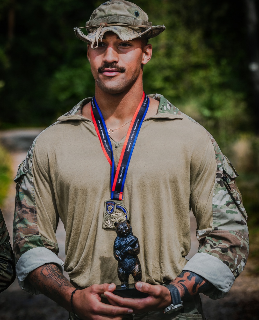 A young male soldier in military camouflage uniform and boonie hat poses holding a bear figurine trophy. He is wearing a medal on a red and blue ribbon lanyard around his neck, suggesting he has won or placed in a military competition. The setting appears to be an outdoor wooded area.