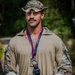 A young male soldier in military camouflage uniform and boonie hat poses holding a bear figurine trophy. He is wearing a medal on a red and blue ribbon lanyard around his neck, suggesting he has won or placed in a military competition. The setting appears to be an outdoor wooded area.