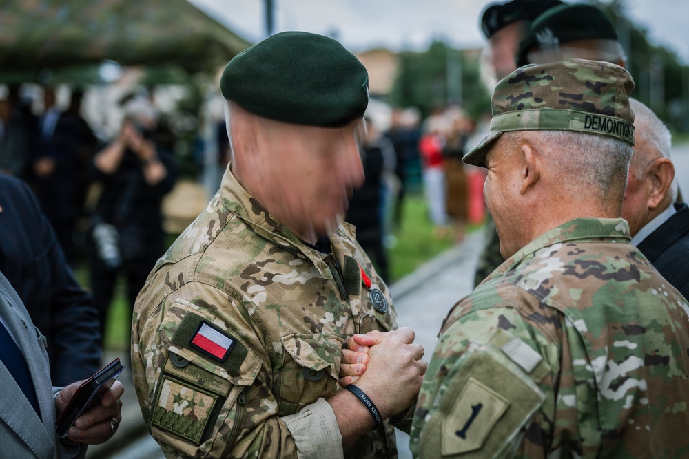 Two military personnel are engaged in what appears to be a medal or decoration ceremony, with one soldier pinning something onto the other's uniform. The soldier on the left wears a Polish Army uniform with a Polish flag patch and green beret, while the soldier on the right wears a US Army OCP uniform with a '1st Infantry Division' patch and the name tape 'DEMONTIGNY'. The event appears to be taking place outdoors at a military installation with other attendees visible in the background.