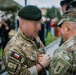 Two military personnel are engaged in what appears to be a medal or decoration ceremony, with one soldier pinning something onto the other's uniform. The soldier on the left wears a Polish Army uniform with a Polish flag patch and green beret, while the soldier on the right wears a US Army OCP uniform with a '1st Infantry Division' patch and the name tape 'DEMONTIGNY'. The event appears to be taking place outdoors at a military installation with other attendees visible in the background.
