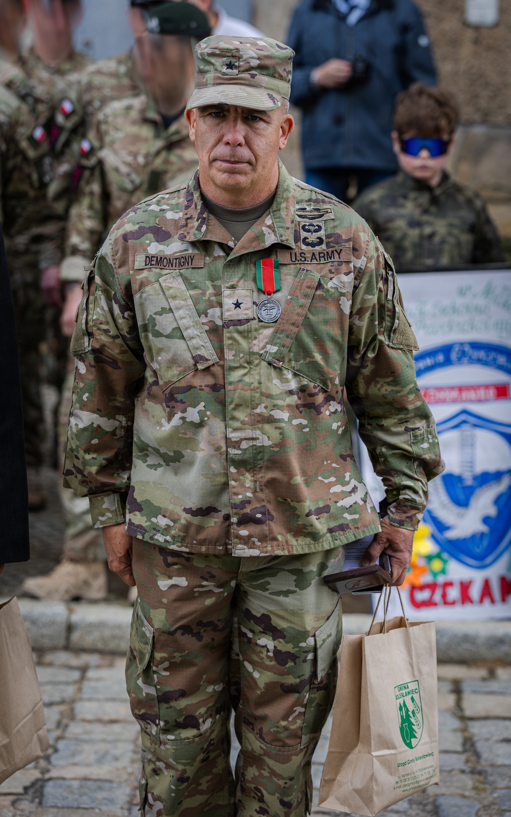 A U.S. Army Brigadier General named Demontigny stands in military OCP (Operational Camouflage Pattern) uniform at what appears to be an outdoor ceremony or event. He is wearing a single-star rank insignia on his chest and cap, along with a medal with a red and green ribbon. He is holding a paper shopping bag and what appears to be a booklet or folder.