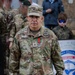 A U.S. Army Brigadier General named Demontigny stands in military OCP (Operational Camouflage Pattern) uniform at what appears to be an outdoor ceremony or event. He is wearing a single-star rank insignia on his chest and cap, along with a medal with a red and green ribbon. He is holding a paper shopping bag and what appears to be a booklet or folder.