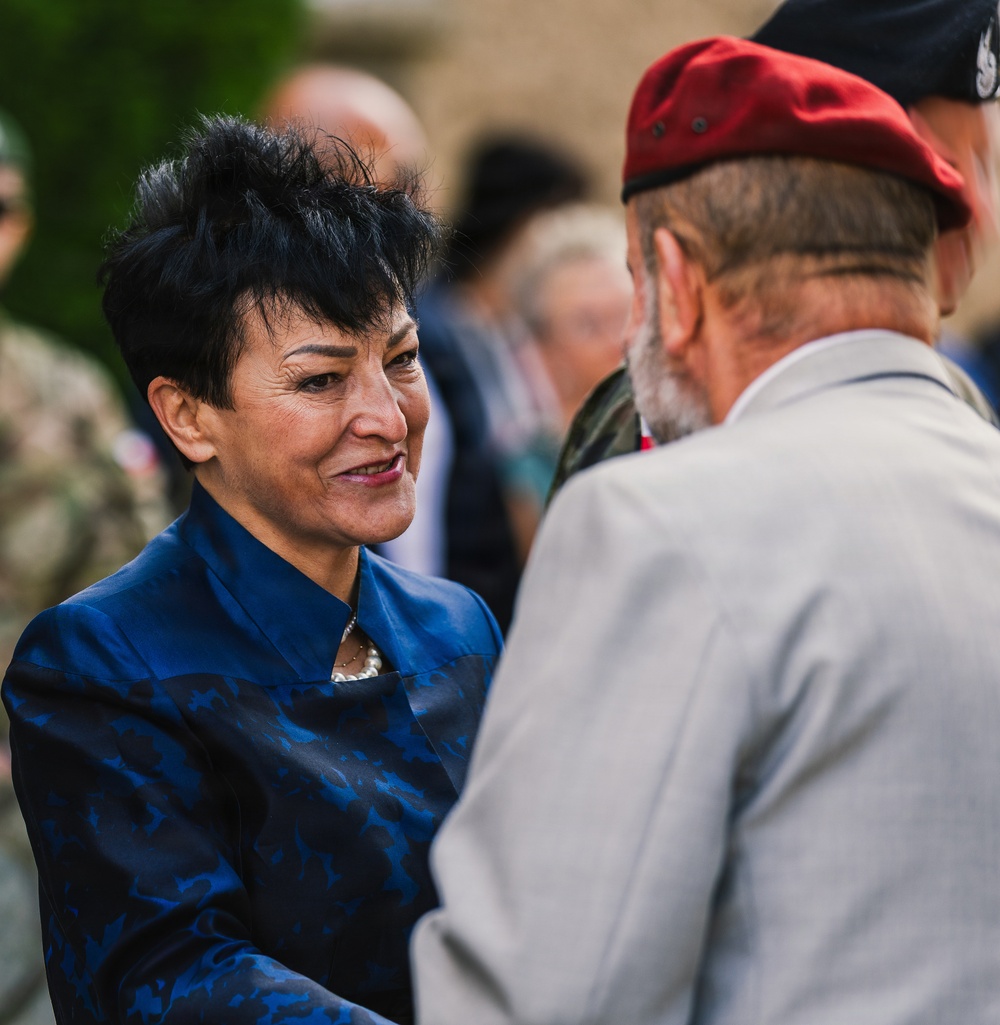 A woman in a blue patterned jacket is engaged in a warm greeting or conversation with an older man wearing a red beret and gray suit jacket. The setting appears to be an outdoor ceremonial or commemorative event, suggested by the presence of military personnel in camouflage uniforms visible in the background. The interaction appears formal yet cordial in nature.