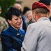 A woman in a blue patterned jacket is engaged in a warm greeting or conversation with an older man wearing a red beret and gray suit jacket. The setting appears to be an outdoor ceremonial or commemorative event, suggested by the presence of military personnel in camouflage uniforms visible in the background. The interaction appears formal yet cordial in nature.